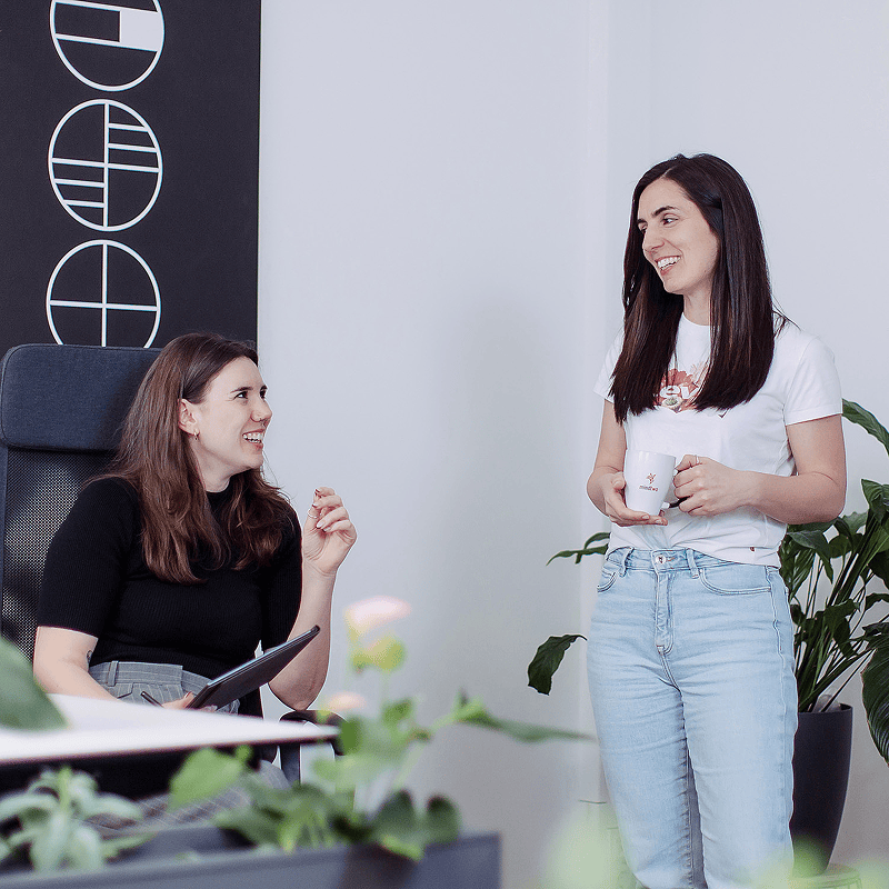 Zwei Frauen unterhalten sich l&auml;chelnd in einem modernen B&uuml;ro mit Pflanzen